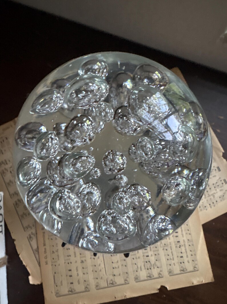 May include: A clear glass paperweight with a bubble design. The round paperweight is filled with numerous spherical air bubbles, creating a textured appearance. The paperweight sits on top of vintage sheet music.
