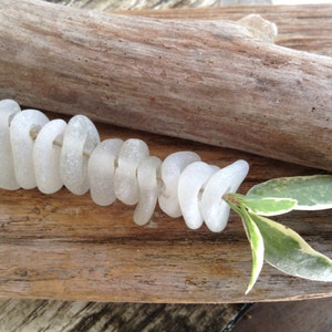 Puede incluir: Una hebra de cuentas de vidrio de mar blanco dispuestas en espiral sobre un trozo de madera a la deriva con una hoja verde.