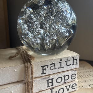 May include: A clear glass paperweight with numerous air bubbles sits atop a stack of three white blocks tied with twine. The blocks display the words "Faith", "Hope", and "Love" in black letters. The background includes sheet music.