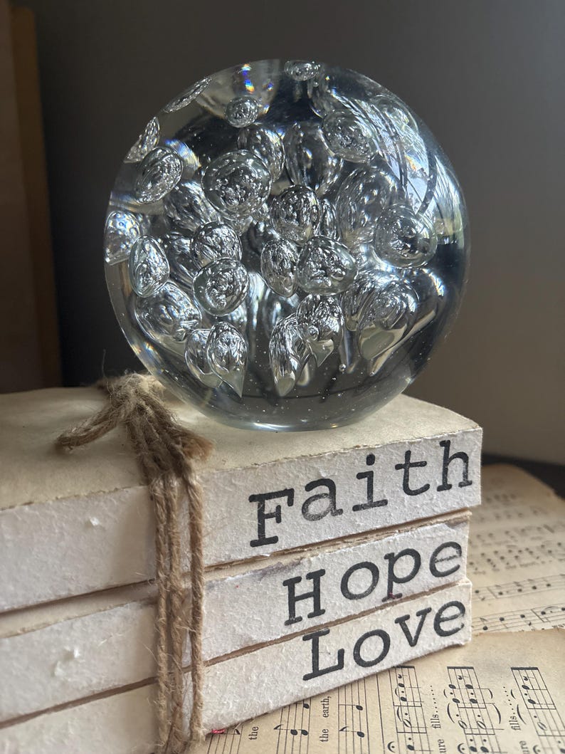 May include: A clear glass paperweight with numerous air bubbles sits atop a stack of three white blocks tied with twine. The blocks display the words "Faith", "Hope", and "Love" in black letters. The background includes sheet music.
