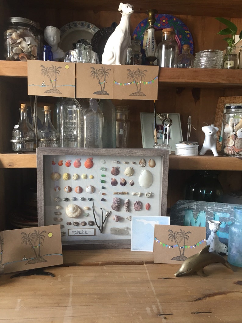 May include: A wooden cabinet displays various items, including seashells, greeting cards with palm tree illustrations, glass bottles, and decorative figurines. A framed collection of seashells is a focal point, alongside a brass dolphin figurine.