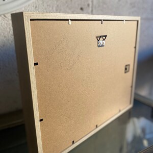 May include: A rectangular wooden picture frame with a light-colored wood finish. The back of the frame is visible, showing a cardboard backing, a metal hanger, and handwritten text. The frame is designed for wall display.