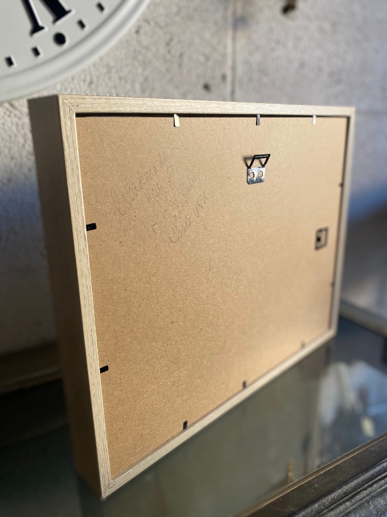 May include: A rectangular wooden picture frame with a light-colored wood finish. The back of the frame is visible, showing a cardboard backing, a metal hanger, and handwritten text. The frame is designed for wall display.