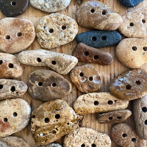 Könnte beinhalten: Eine Sammlung von Naturstein-Knöpfen in verschiedenen Formen und Farben, darunter Braun, Beige und Grau. Jeder Knopf hat zwei gleichmäßig verteilte Löcher zum Annähen. Die Knöpfe sind auf einer Holzoberfläche angeordnet.