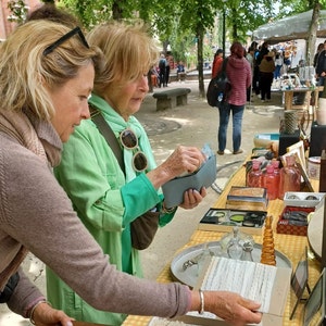 May include: Two women are looking at a blue wallet. The woman on the left is wearing a brown sweater and the woman on the right is wearing a green sweater. They are standing in front of a table with various items on it, including a glass bottle, a pink bottle, and a small box.