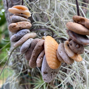 Puede incluir: Un collar hecho de piedras naturales y conchas. Las piedras son de varios tonos de marrón y gris, y las conchas son de color beige claro. El collar está encordado en un cordón marrón.
