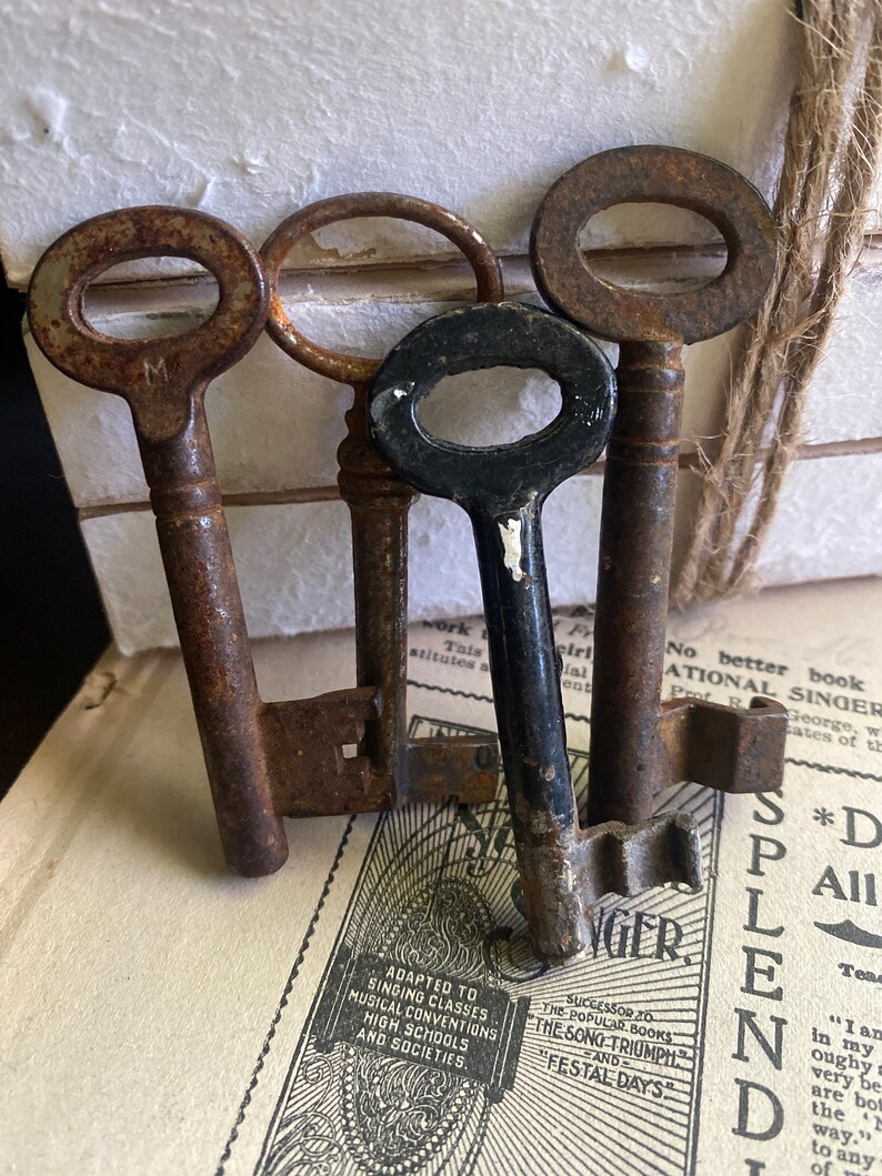 May include: Three antique keys, one black and two rusty brown, with a single keyhole on each. The keys are arranged on top of a vintage book with text about singing classes, musical conventions, high schools, and societies.