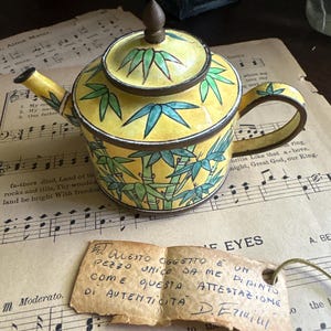 Op de afbeelding: Een gele theepot met een donker metalen rand, deksel en handvat, versierd met groene bamboebladpatronen. Een klein, rechthoekig label van verouderd papier is bevestigd, voorzien van handgeschreven Italiaanse tekst en een handtekening.