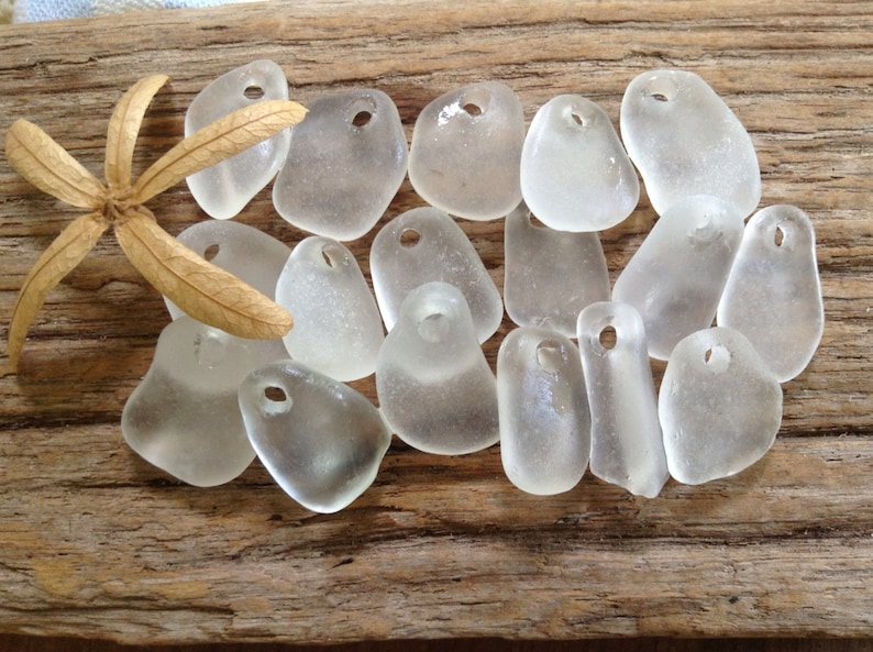 Puede incluir: Una colecci&oacute;n de 15 colgantes de vidrio de mar liso y transparente con agujeros para encordar. Los colgantes son de varias formas y tama&ntilde;os, y est&aacute;n dispuestos sobre un trozo de madera desgastada.