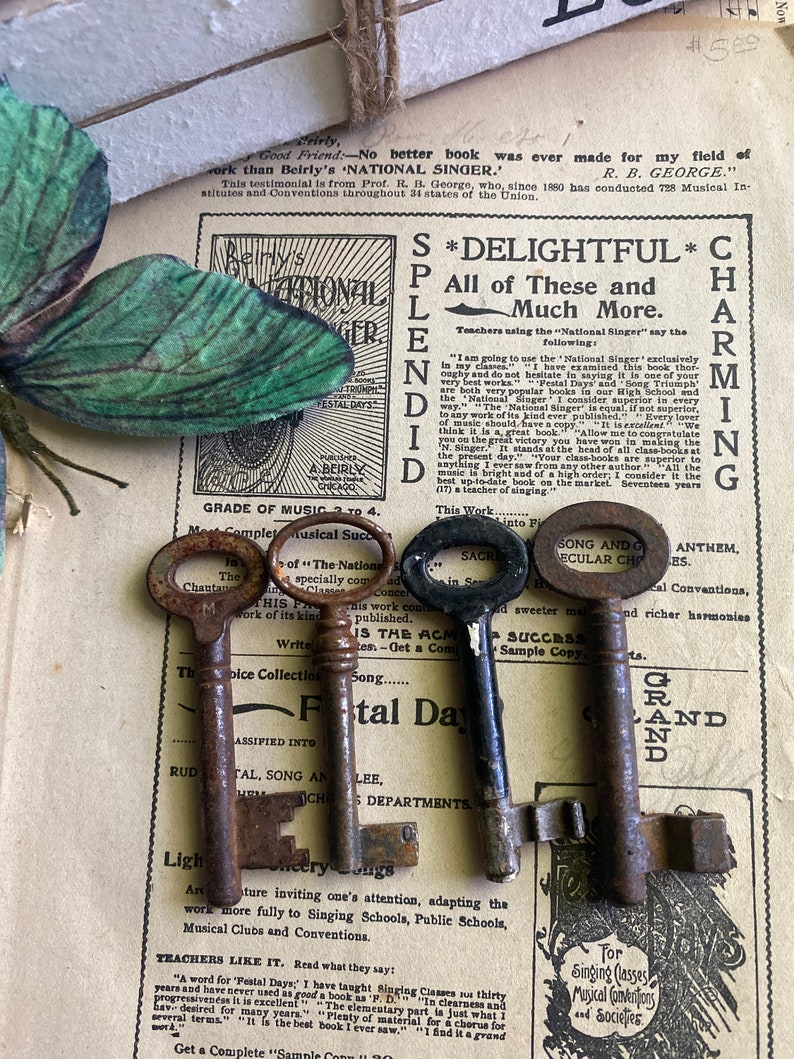 May include: Three antique keys with tarnished metal finishes. The keys are arranged on a page of a vintage music book with text about a "National Singer" and "Festal Days".
