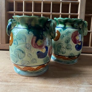 Puede incluir: Dos jarrones de cerámica con fondo azul claro y diseños florales en naranja, rojo y azul. Los jarrones tienen un borde y asas verdes. La base de los jarrones tiene una banda naranja y azul.