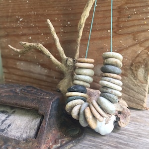 May include: A necklace made of stacked, smooth, round stones in shades of gray, brown, and white. The stones are strung together with a thin blue cord and hang from a branch.