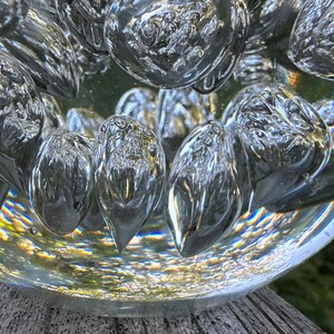May include: Close-up of a clear glass decorative object with bubble-like shapes. The glass has a reflective quality, capturing light and creating a shimmering effect. The object sits on a weathered wooden surface.