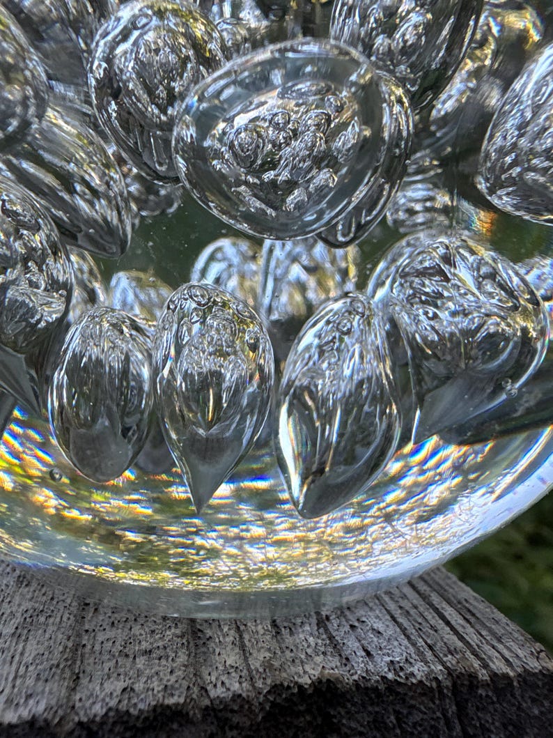 May include: Close-up of a clear glass decorative object with bubble-like shapes. The glass has a reflective quality, capturing light and creating a shimmering effect. The object sits on a weathered wooden surface.
