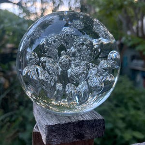 May include: A clear glass sphere with numerous air bubbles suspended within. The sphere is resting on a weathered wooden post, with a blurred green background of foliage. The bubbles create a textured, refractive effect.