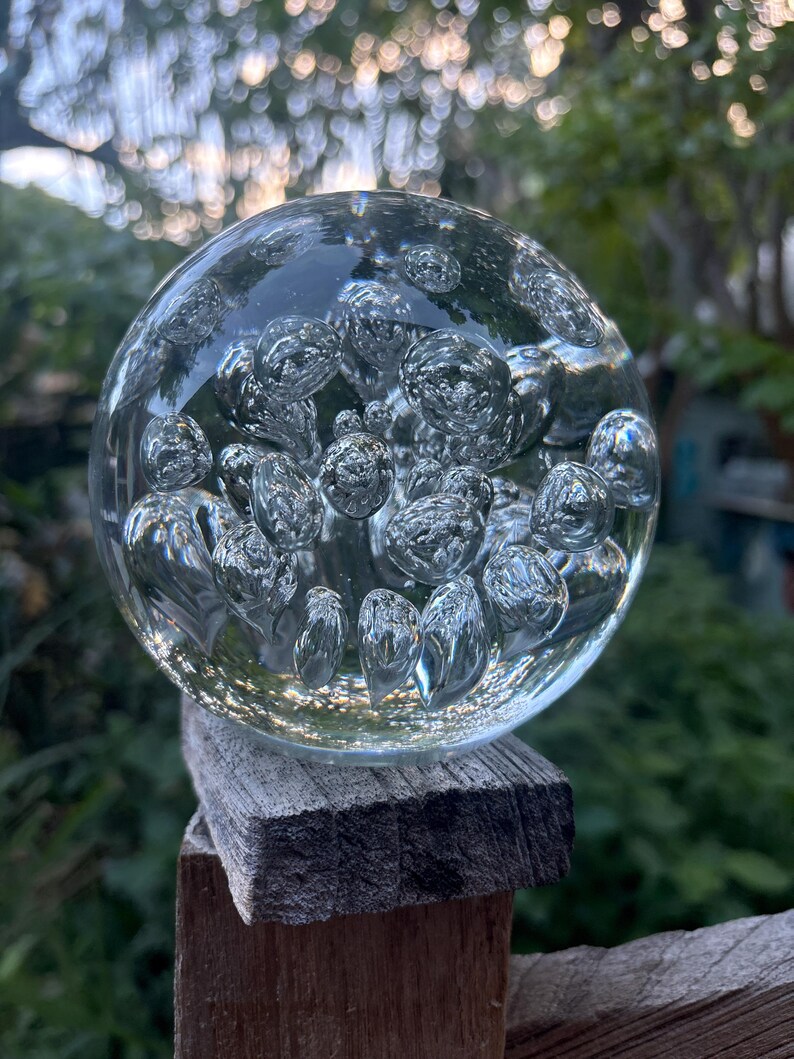 May include: A clear glass sphere with numerous air bubbles suspended within. The sphere is resting on a weathered wooden post, with a blurred green background of foliage. The bubbles create a textured, refractive effect.