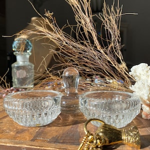 May include: A still life arrangement featuring two small, clear glass bowls with a textured design, a clear glass stopper, and a gold-colored key set. Dried twigs and a decorative bottle are in the background.