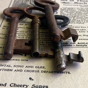 May include: Three antique keys made of metal with a worn, aged finish. The keys are arranged on top of a vintage sheet of paper with text that reads "S*DELIGHT P All of These Much".