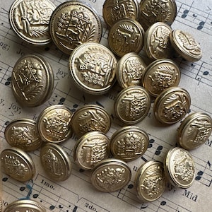 May include: A collection of gold-colored metal buttons with intricate embossed designs. The buttons vary in size and feature detailed crests, emblems, and floral patterns. They are scattered on a sheet of vintage sheet music.