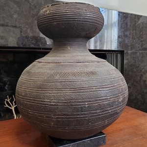 Puede incluir: Un jarrón de cerámica marrón oscuro, hecho a mano, con un cuerpo bulboso y un cuello más pequeño y cilíndrico. El jarrón presenta líneas grabadas horizontales y diagonales, y se apoya sobre una base cuadrada negra. El jarrón tiene una superficie texturizada.