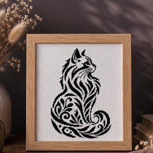 Könnte beinhalten: Ein gerahmtes Kreuzstichbild einer stilisierten schwarzen Katze mit komplizierten floralen und wirbelnden Mustern. Die Katze ist vor einem weißen Hintergrund in einem hellbraunen Holzrahmen dargestellt. Das Kunstwerk wird auf einer Holzoberfläche mit dekorativen Elementen präsentiert.