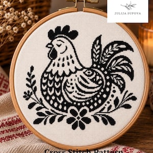 Peut inclure: Un tambour à broder circulaire en bois contenant un panneau en tissu blanc avec un motif de point de croix noir représentant un coq entouré d'éléments floraux. Le texte JULIJA SUPOVA est visible en haut à droite et Cross Stitch Pattern en bas.