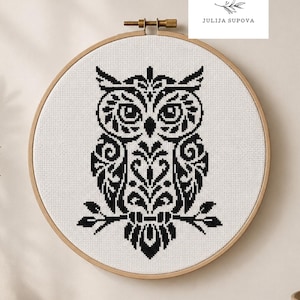 Peut inclure: Un motif de broderie au point de croix noir et blanc représentant un hibou sur tissu blanc, encadré dans un cercle à broder en bois. Le hibou est perché sur une branche. Le texte "Cozy Cross Stitch Pattern" est en bas.