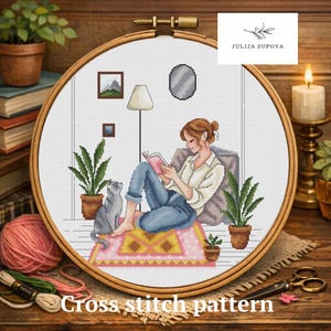 以下が含まれることがあります： 木製のフレームに入ったクロスステッチのデザイン。本を読む人、灰色の猫、植物、ランプが描かれています。「Cross stitch pattern」の文字が下にあります。ピンクのラグと居心地の良いインテリアシーンが特徴です。