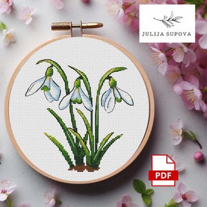 May include: A cross-stitch embroidery of snowdrop flowers in a wooden hoop. The design features white and green flowers with long green stems and leaves. The hoop is set against a white background with pink flowers.