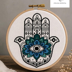 May include: A cross-stitch pattern of a Hamsa hand with an eye in the center, displayed in a wooden embroidery hoop. The design features black and white details with teal and blue accents. The text "Cross Stitch Pattern" is at the bottom.
