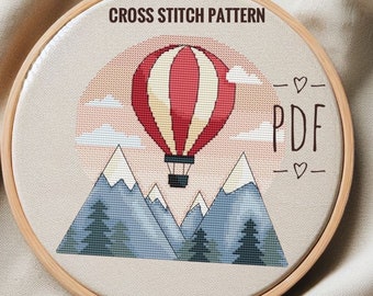 May include: Cross stitch pattern PDF for a hot air balloon flying over mountains with trees. The balloon is red and white with a basket. The mountains are blue and white with snow on the peaks. The trees are green. The background is a light pink.