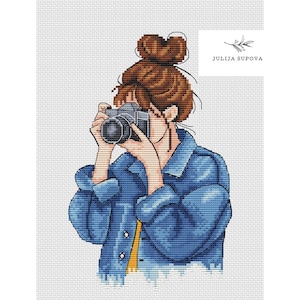 May include: A cross-stitch pattern of a woman with brown hair in a bun, wearing a blue denim jacket and holding a camera. The text "JULIJA SUPOVA" is in the top right corner of the image.