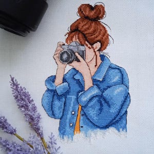 May include: Cross-stitch embroidery of a person with brown hair in a bun wearing a blue denim jacket and holding a camera.