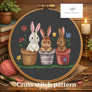 Könnte beinhalten: Kreuzstichmuster mit drei Hasen in Blumentöpfen. Die Hasen sind weiß, braun und hellbraun, mit rosa gefütterten Ohren. Jeder Hase sitzt in einem unterschiedlich farbigen Topf mit Blumen. Der Text "Cross stitch pattern" steht unten.