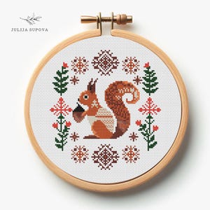 May include: Cross stitch pattern featuring a brown squirrel holding a nut, surrounded by green leaves, red flowers, and brown snowflakes. The pattern is framed in a wooden hoop.