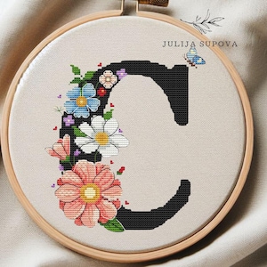 May include: Cross-stitch embroidery of the letter "C" decorated with colorful flowers and a butterfly. The embroidery is set in a wooden hoop, with the text "JULIJA SUPOVA" visible.