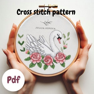 Pode incluir: Padrão de ponto cruz com um cisne branco com rosas cor-de-rosa e folhas verdes. O padrão está em um aro de bordado de madeira e o texto "JULIJA SUPOVA" é visível. O texto "Pdf" também é visível em um círculo rosa.