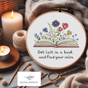 Op de afbeelding: Een ronde houten borduurring met een kruissteekontwerp van een open boek met kleurrijke bloemen die uit de pagina's komen. De tekst "Get lost in a book and find your calm" is in zwarte draad onder de illustratie van het boek geborduurd.