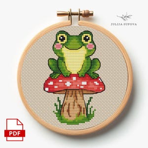 May include: A cross-stitch embroidery hoop featuring a green frog sitting on a red mushroom with white spots. The frog has pink cheeks and yellow belly. The embroidery is set in a wooden hoop. The text "JULIJA SUPOVA" is visible.
