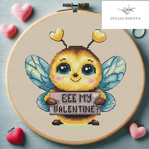 Puede incluir: Un patrón de punto de cruz con una linda abeja de dibujos animados sosteniendo un cartel que dice "Bee My Valentine". La abeja es amarilla y negra con grandes ojos azules y una nariz rosa. El cartel es rosa con letras blancas. El patrón está rodeado por un aro de bordado de madera.