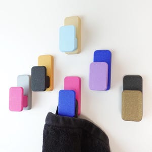 Könnte beinhalten: Eine Sammlung farbenfroher Wandhaken in verschiedenen Farbtönen, darunter Rosa, Blau, Gold und Schwarz. Die Haken sind rechteckig mit abgerundeter Oberseite und an einer weißen Wand befestigt. Ein Haken hält ein schwarzes Kleidungsstück.