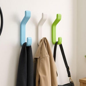 Könnte beinhalten: Drei Wandhaken in hellblau, weiß und grün. Die Haken sind J-förmig und an einer weißen Wand montiert. Ein schwarzer Schal, eine braune Jacke und eine schwarze Handtasche hängen an den Haken.