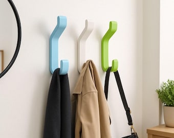 May include: Three wall hooks in light blue, white, and green. The hooks are shaped like a 'J' and are mounted on a white wall. A black scarf, a brown jacket, and a black handbag are hanging on the hooks.