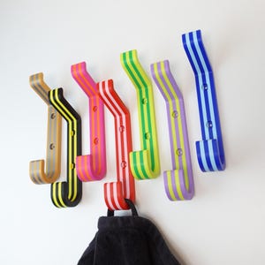 Könnte beinhalten: Ein Set aus sechs Wandhaken in verschiedenen Farben, darunter Beige, Schwarz, Pink, Rot, Limettengrün, Lila und Blau. Jeder Haken hat ein Streifenmuster und zwei kreisförmige Schraublöcher zur Befestigung. Ein schwarzes Handtuch hängt am roten Haken.