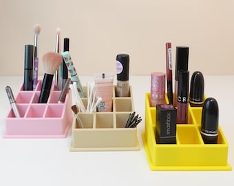 Könnte beinhalten: Drei farbige Kosmetik-Organizer in Rosa, Beige und Gelb, gefüllt mit Make-up-Pinseln, Lippenstiften und anderen Schönheitsprodukten. Jeder Organizer hat mehrere Fächer. Eine Vielzahl von Kosmetikartikeln ist sichtbar, darunter Lippenstifte und Pinsel.
