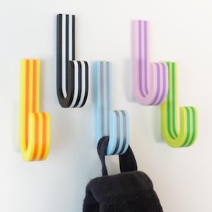 May include: Five brightly coloured, striped wall hooks in yellow, black, blue, pink, and green. Each hook is a curved 'J' shape, mounted on a white wall. A black item hangs from the blue hook.