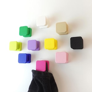 May include: A selection of brightly coloured square wall hooks in shades of green, white, yellow, purple, blue, pink, and black. One hook is holding a black towel. The hooks are mounted on a white wall, providing a modern storage solution.