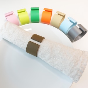 Può includere: Una tovagliolo di lino bianco arrotolato su un piatto bianco, tenuto da un portatovagliolo marrone. Sei portatovaglioli colorati in rosa, verde-giallo, verde, arancione, beige, azzurro e grigio sono disposti attorno al tovagliolo.