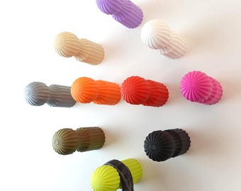 May include: A selection of vibrantly coloured wall hooks in shades of purple, white, beige, grey, orange, red, pink, green, brown, and black. The hooks feature a textured, ribbed design and are mounted on a white wall. A scarf hangs from a green hook.