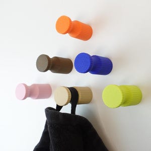 May include: A selection of brightly coloured wall hooks in orange, brown, blue, pink, beige, and green. A black towel hangs from one hook. The hooks are mounted on a white wall, providing a modern storage solution.
